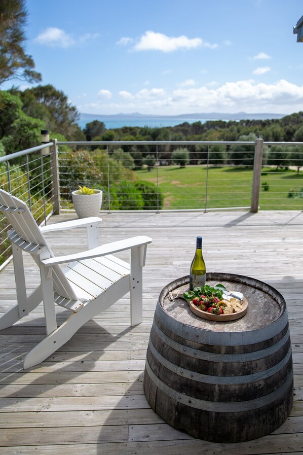 Una terraza de madera con una silla blanca, un barril rústico y una botella de vino, con vistas a un paisaje verde.