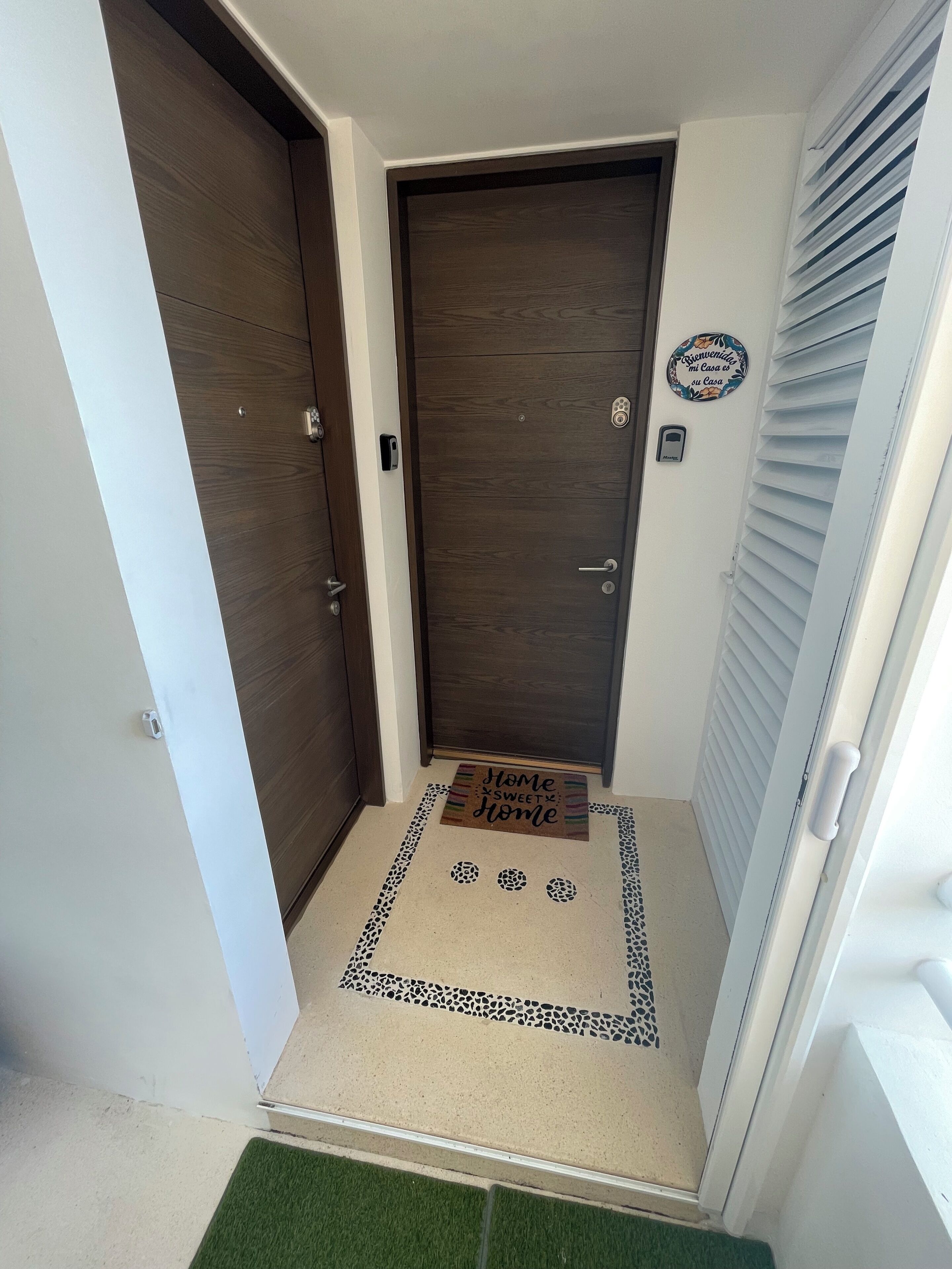 A hallway with two dark wooden doors, a welcome mat with 'Home Sweet Home' text, and a wall-mounted sign.