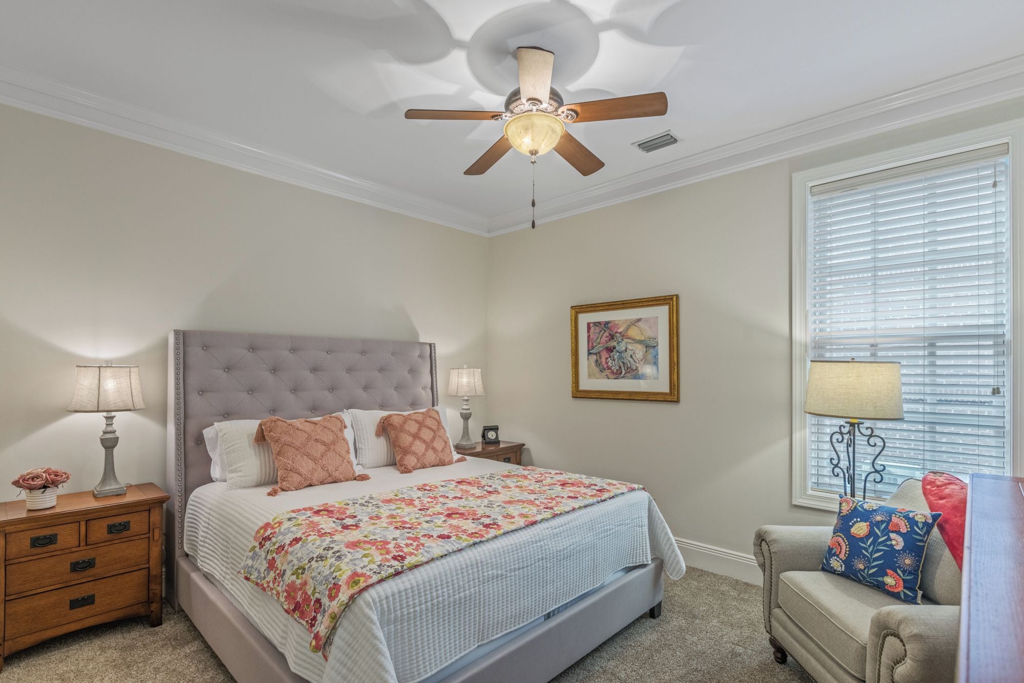 A bedroom with a tufted headboard, a bed with floral bedding, a nightstand with lamps, a window with blinds, and a ceiling fan.