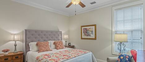 A bedroom with a tufted headboard, a bed with floral bedding, a nightstand with lamps, a window with blinds, and a ceiling fan.