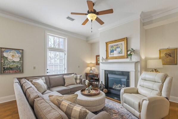 A living room with a sofa, armchair, fireplace, ceiling fan, and artwork on the walls.