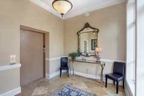 A hallway with a door, a mirror, two chairs, a console table with a lamp, and a patterned rug.