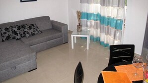 Un salon avec un canapé gris, une petite table blanche et un coin repas comprenant des chaises et une bouteille de vin.