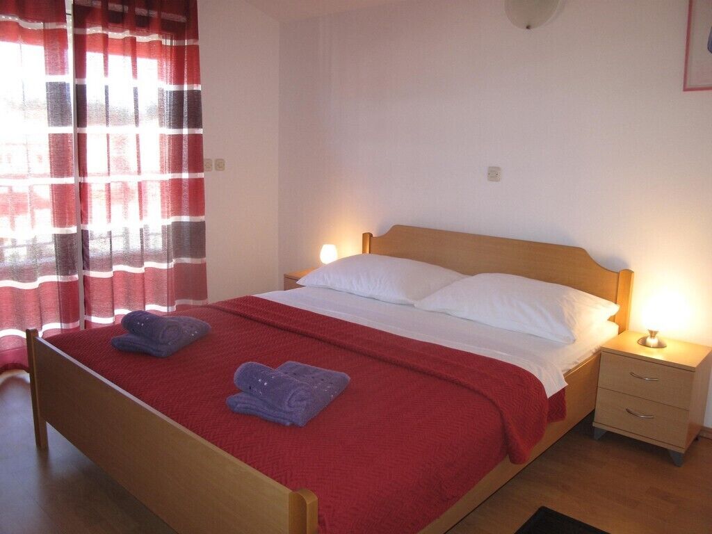 A single bed with a red bedspread and white pillows, two bedside tables with lamps, and a window with red curtains.