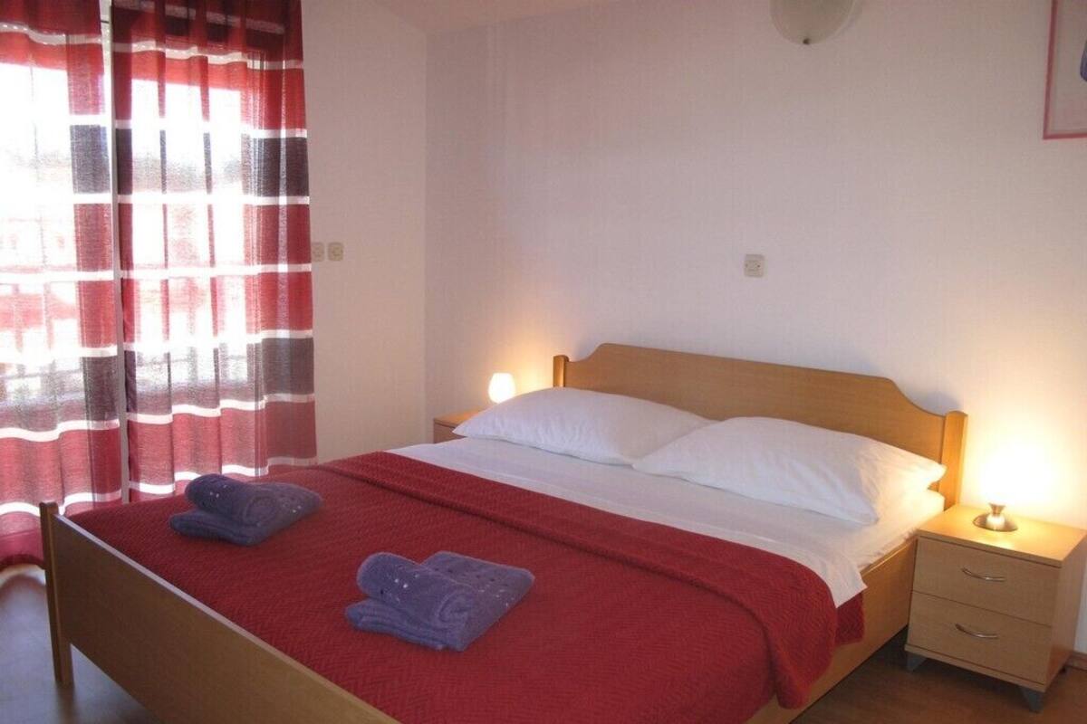 A single bed with a red bedspread and white pillows, two bedside tables with lamps, and a window with red curtains.