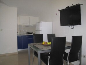 A compact kitchen with a dining table and chairs, a flat-screen TV mounted on the wall, and a refrigerator.