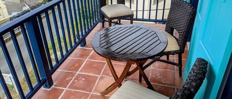 Un balcon avec une table ronde et deux chaises en osier, une rambarde bleue et une vue sur la rue en contrebas.