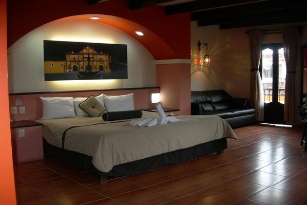 A hotel room with a bed, a black leather sofa, a painting on the wall, and a window with curtains. - Catedral Hotel (San Cristóbal de las Casas)