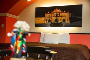 A hotel room with a bed, pillows, a painting of a building, and a colorful sculpture on a table. - Catedral Hotel (San Cristóbal de las Casas)