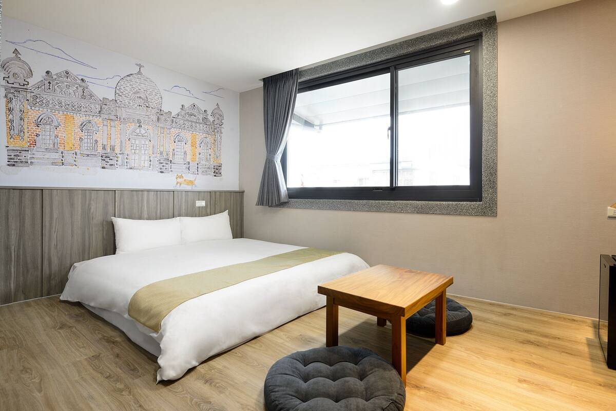 A bedroom with a bed, a small table, a grey ottoman, a window with curtains, and a wall mural.