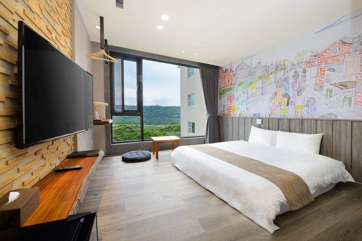 A modern hotel room with a large bed, a flat-screen TV, a wooden wall, and a large window offering a view of greenery.