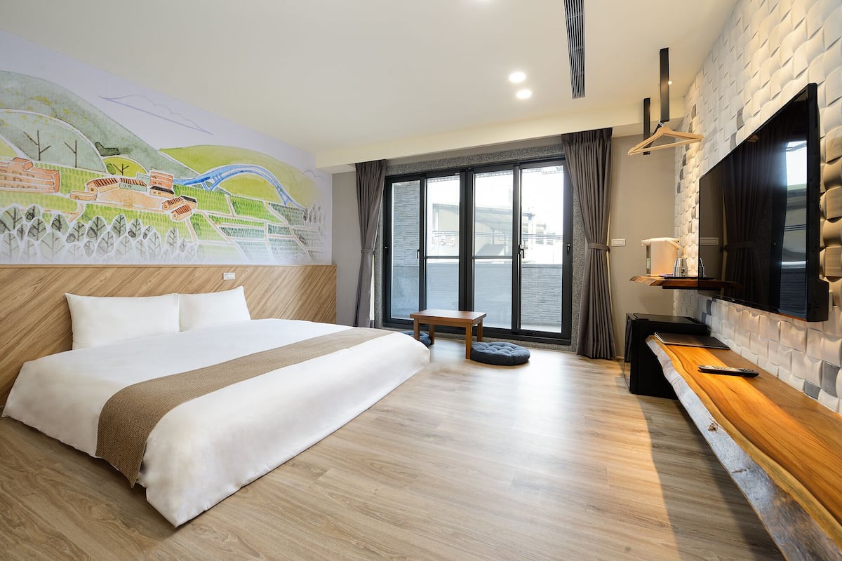 A hotel room with a large bed, a wooden headboard, a map mural, a wooden bench, a TV, and a large window with a view of a building.