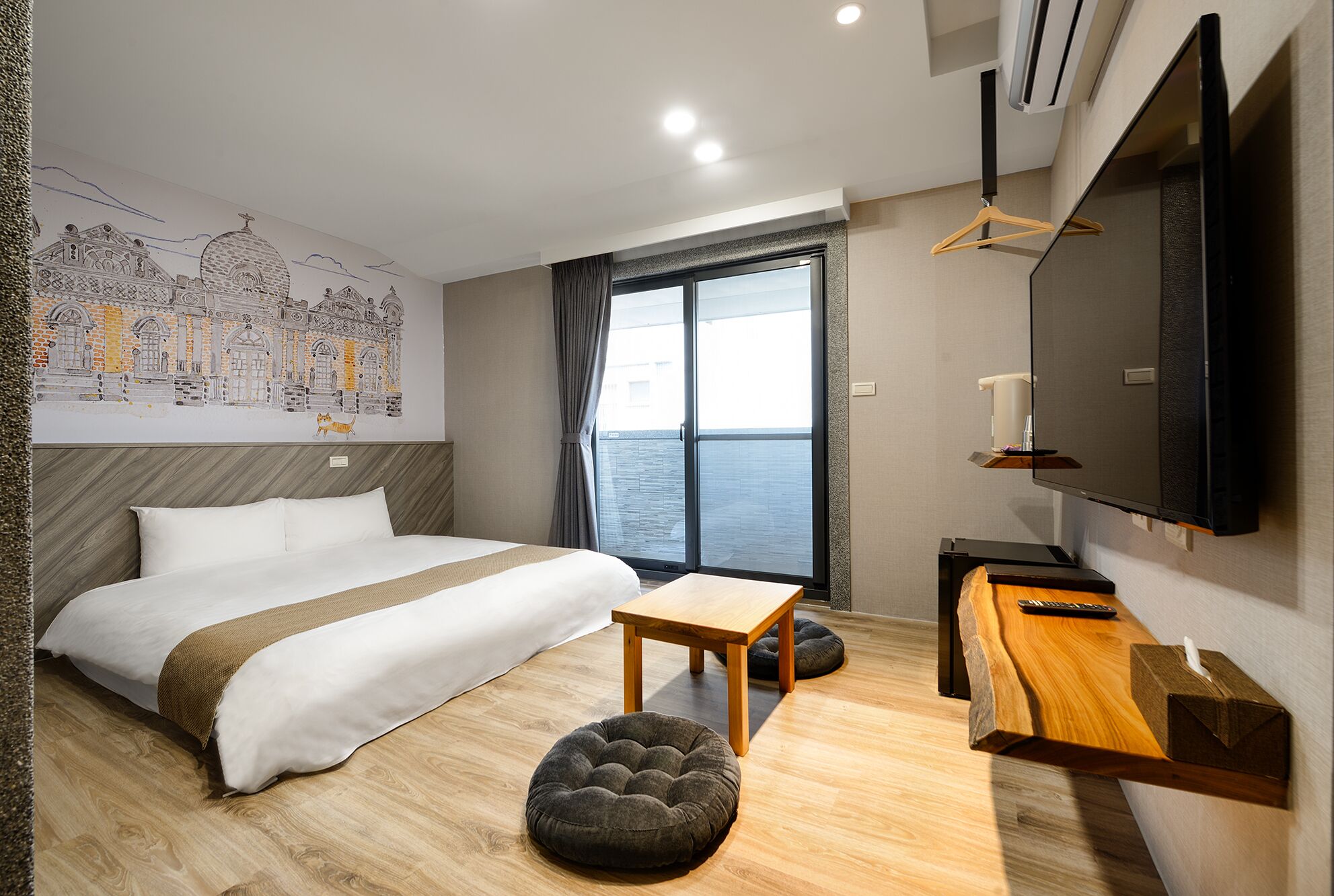 Ein modernes Hotelzimmer mit einem großen Bett, einem Holztisch, einem Flachbildfernseher und einem Wandgemälde mit Stadtansicht.