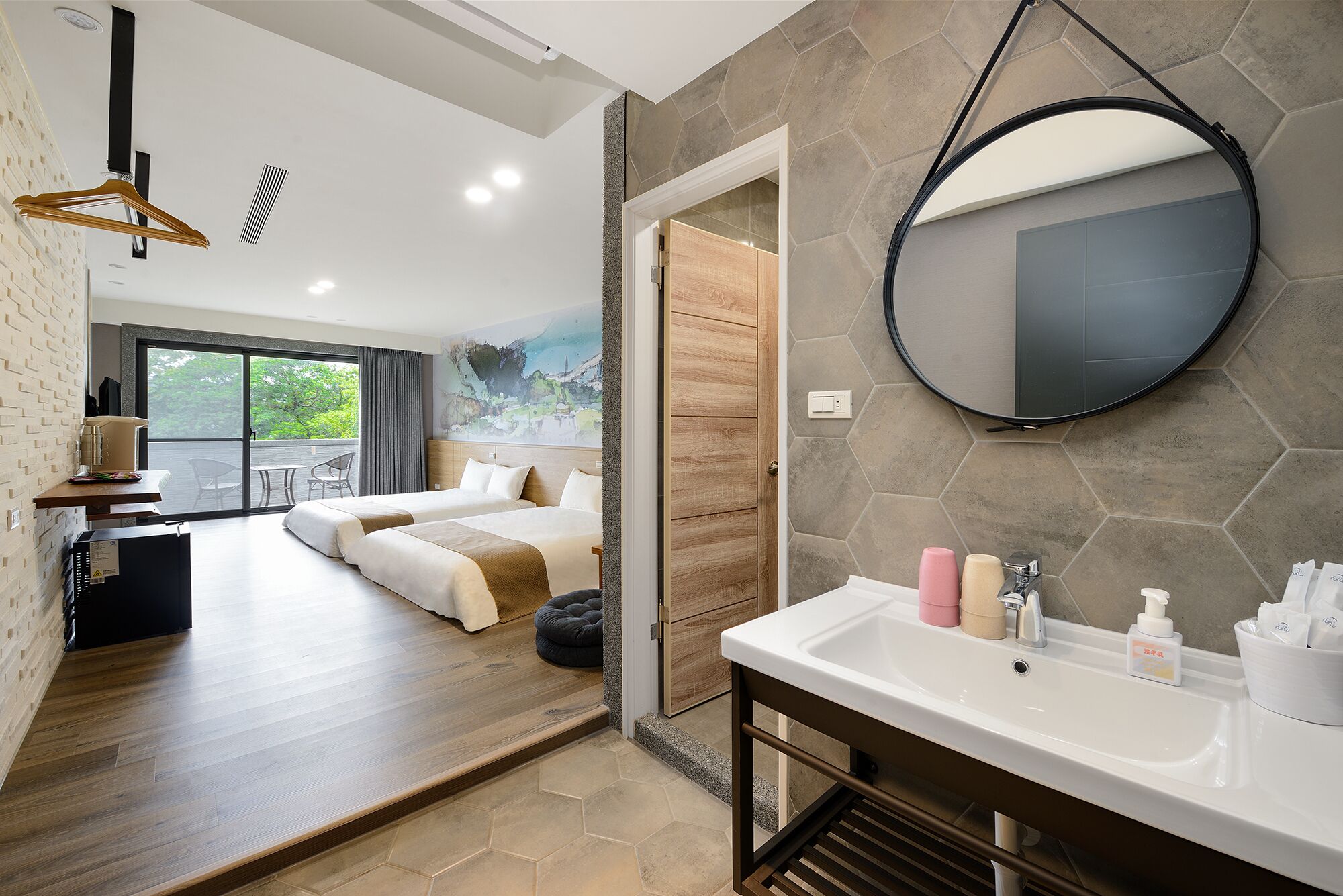Ein modernes Hotelzimmer mit einem großen Bett, einem runden Spiegel, einem Badezimmer mit Waschbecken und Pflegeprodukten sowie Blick ins Freie.