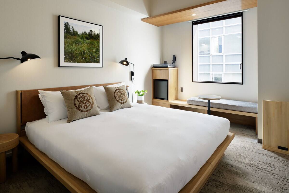 Una habitación de hotel moderna con una cama grande, un escritorio, una silla, una mesita, un televisor y un ventanal amplio.