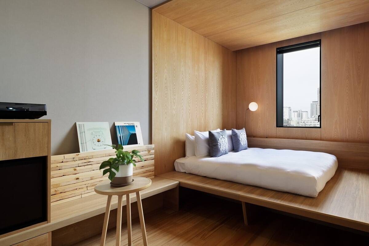 Habitación compacta con cama, una pequeña mesa con planta, televisor de pared y ventana con vista a la ciudad.