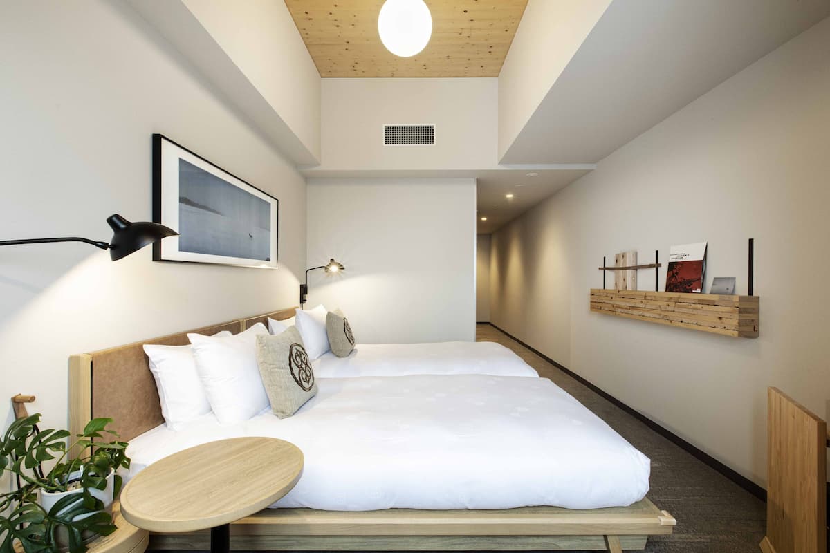 Una habitación de hotel moderna con una cama grande, mesitas de noche, una pequeña mesa redonda, un estante en la pared con libros y una lámpara en el techo.