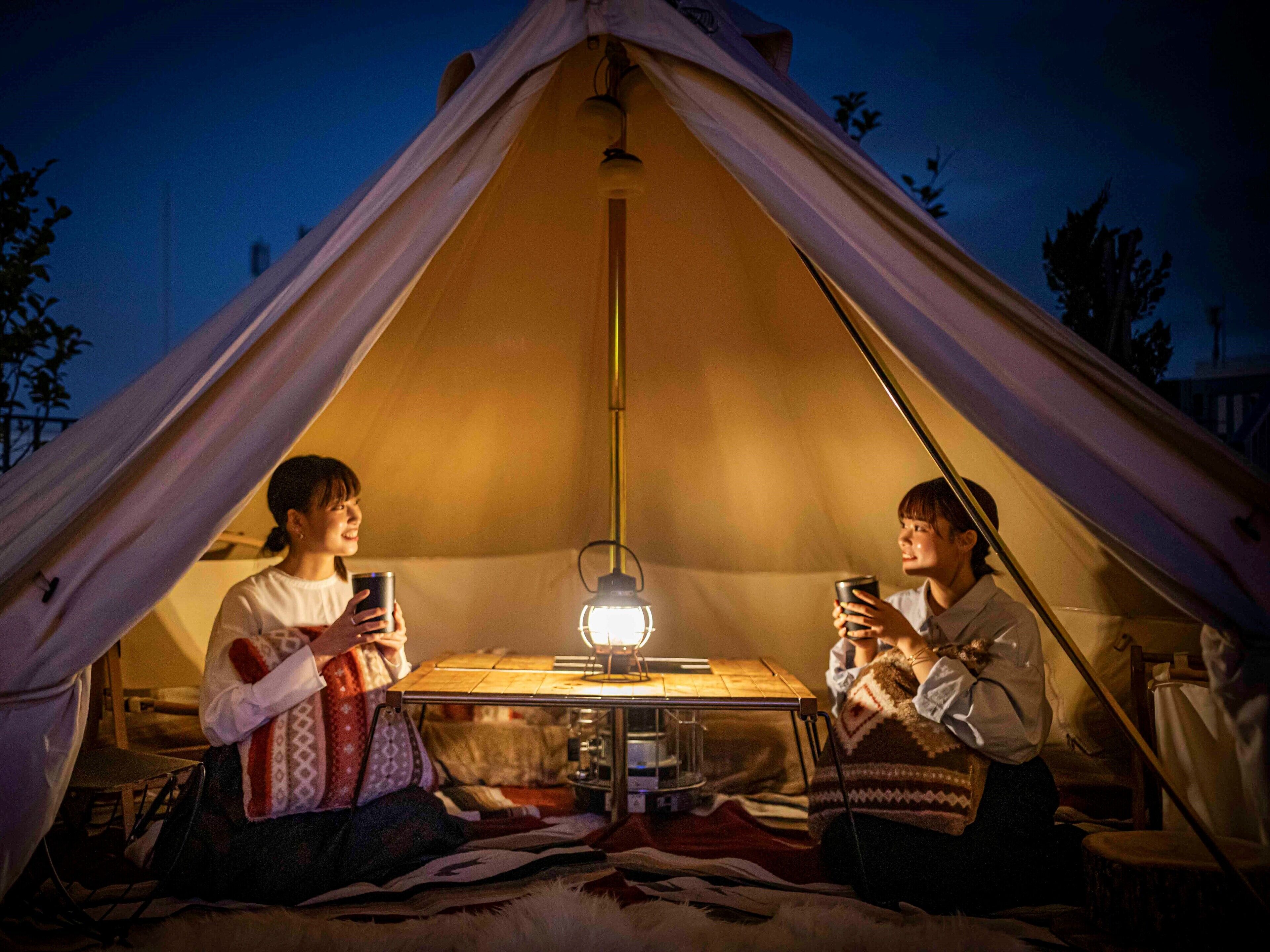 Zwei Personen sitzen in einem Zelt, eine Tisch und eine Lampe stehen zwischen ihnen.