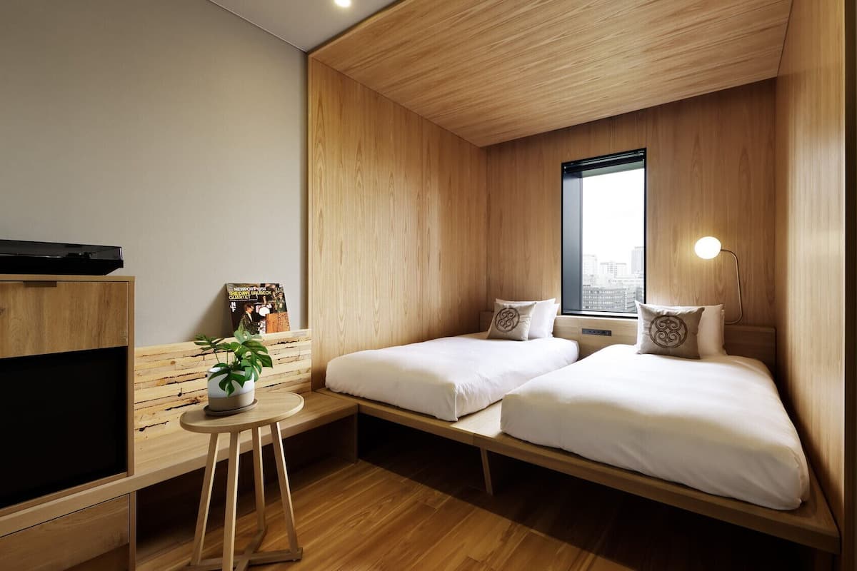 Une chambre d’hôtel compacte, équipée d’une télévision, d’une petite table avec une plante, de deux lits, d’une fenêtre et d’une lampe de chevet.