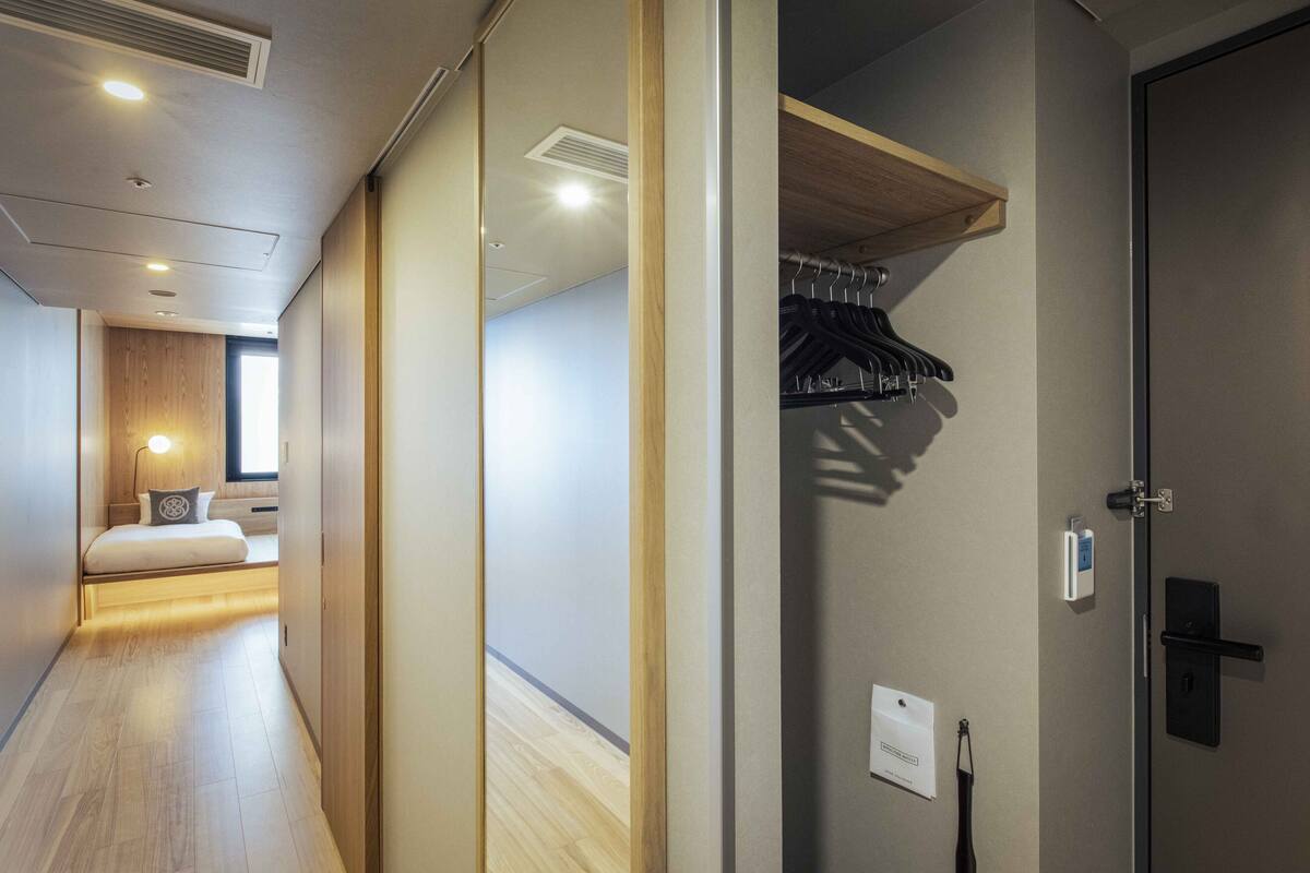 Una habitación de hotel con piso de madera, una cama con almohada, una mesita de noche, un calentador en la pared y una puerta con perchero.