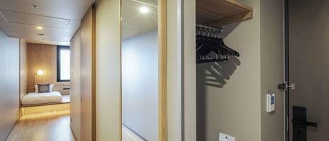 A hotel room with a wooden floor, a bed with a pillow, a bedside table, a wall-mounted heater, and a door with a coat rack.