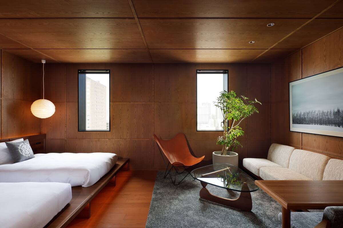 Una habitación de hotel moderna con paredes de madera, un ventanal grande, un sofá, una silla, una mesa de centro de vidrio y una cama con ropa blanca.
