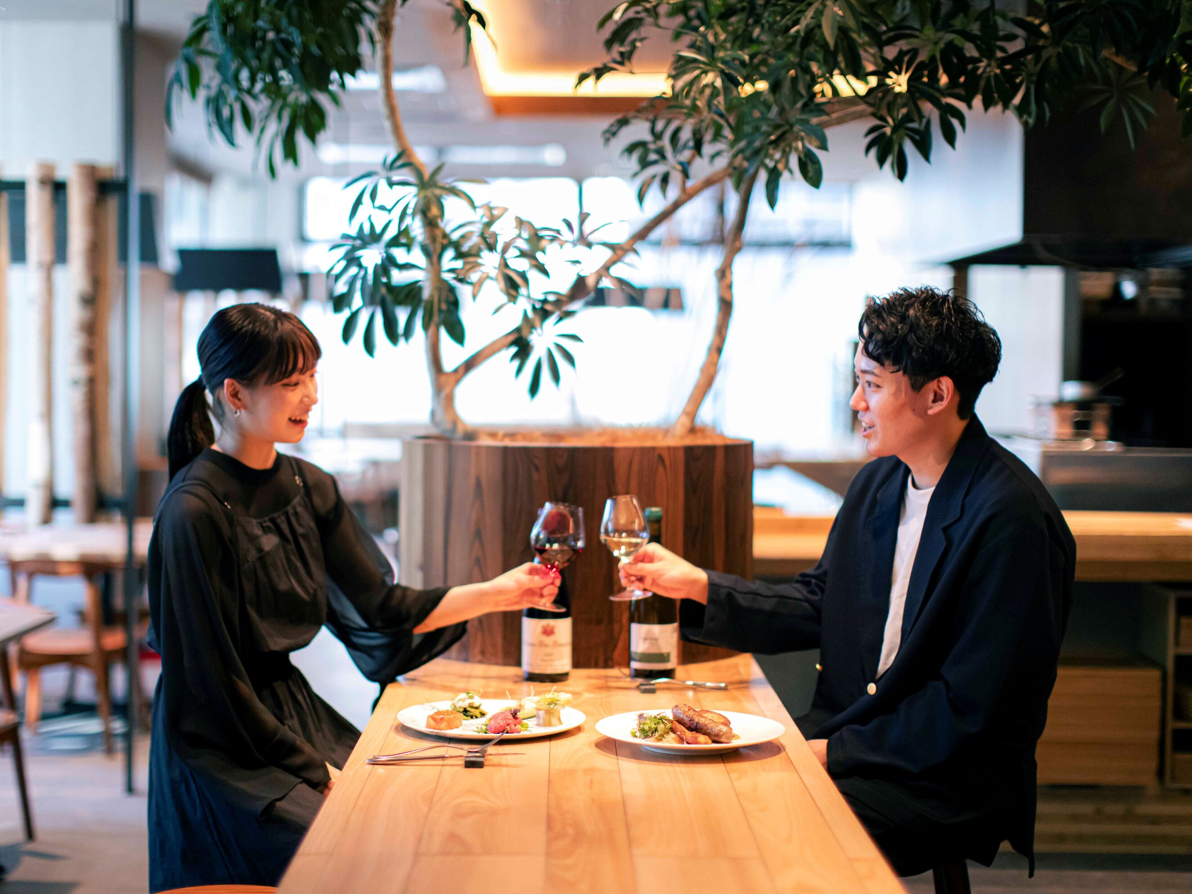 A couple toasting with wine glasses at a wooden table with plates of food, surrounded by indoor plants and a modern kitchen in the background.