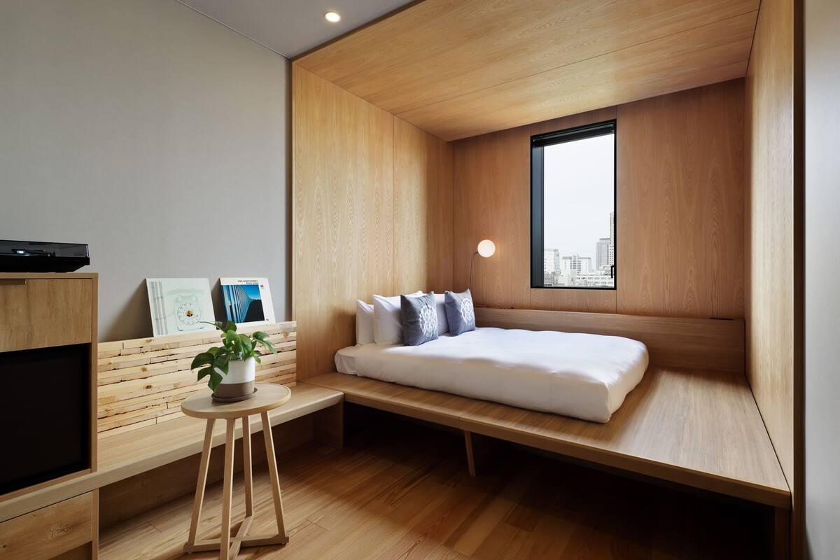 Una habitación moderna con una cama, una mesita con una planta y un televisor sobre un mueble de madera.