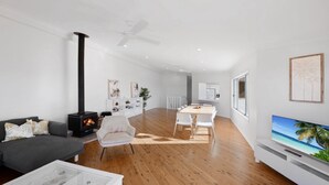 A modern living room with a fireplace, a dining area, and a TV displaying a beach scene. - Ocean Vue - Stunning Views! (AVOCA BEACH)