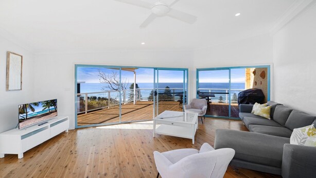 A spacious living room with a large TV, a grey sofa, a white coffee table, and a balcony with a view of the sea. - Ocean Vue - Stunning Views! (AVOCA BEACH)