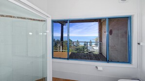 A bathroom with a glass shower enclosure, a window with a view of the sea, and a wooden deck outside. - Ocean Vue - Stunning Views! (AVOCA BEACH)