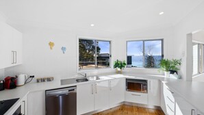 A modern kitchen with white cabinets, stainless steel appliances, and large windows offering a view of the sea. - Ocean Vue - Stunning Views! (AVOCA BEACH)