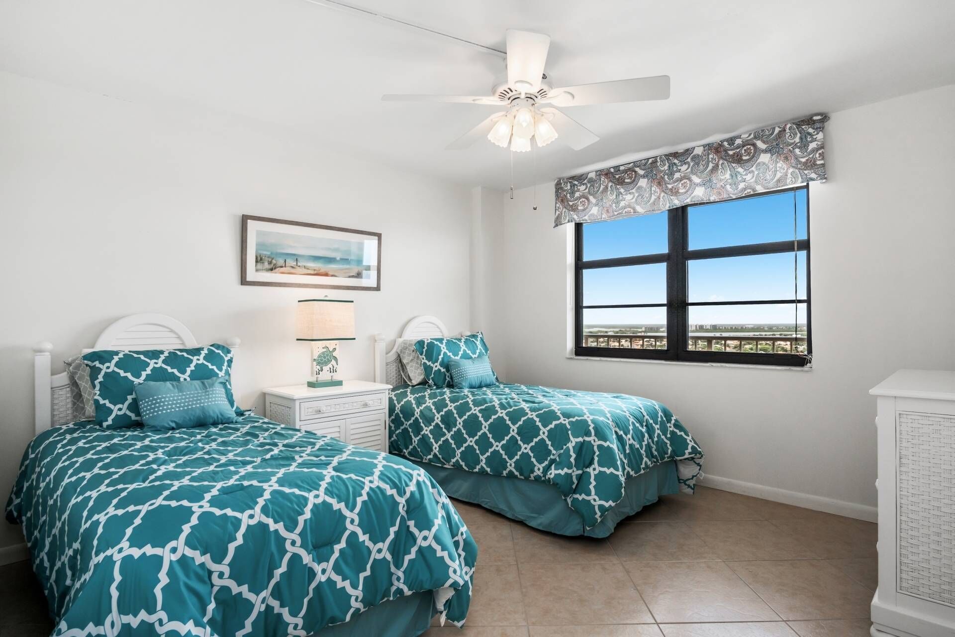 A bedroom with two beds, a ceiling fan, a window with a view, and a nightstand with a lamp.