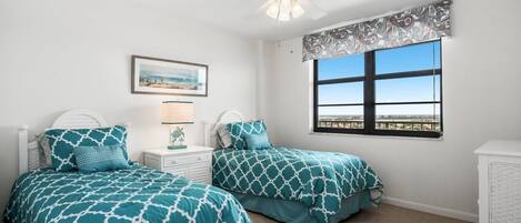 A bedroom with two beds, a ceiling fan, a window with a view, and a nightstand with a lamp.