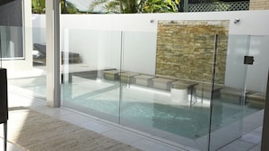 A modern outdoor swimming pool with glass walls, a stone wall, and a seating area with a bench and a table. - Nikatara - Mission Beach (Mission Beach)
