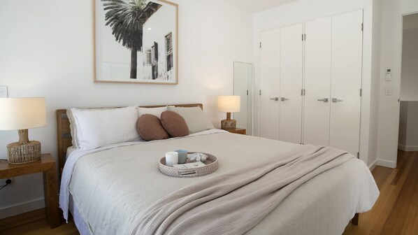 A neatly made bed with a headboard, two bedside lamps, a bedside table, a wall-mounted mirror, and a door with a handle.