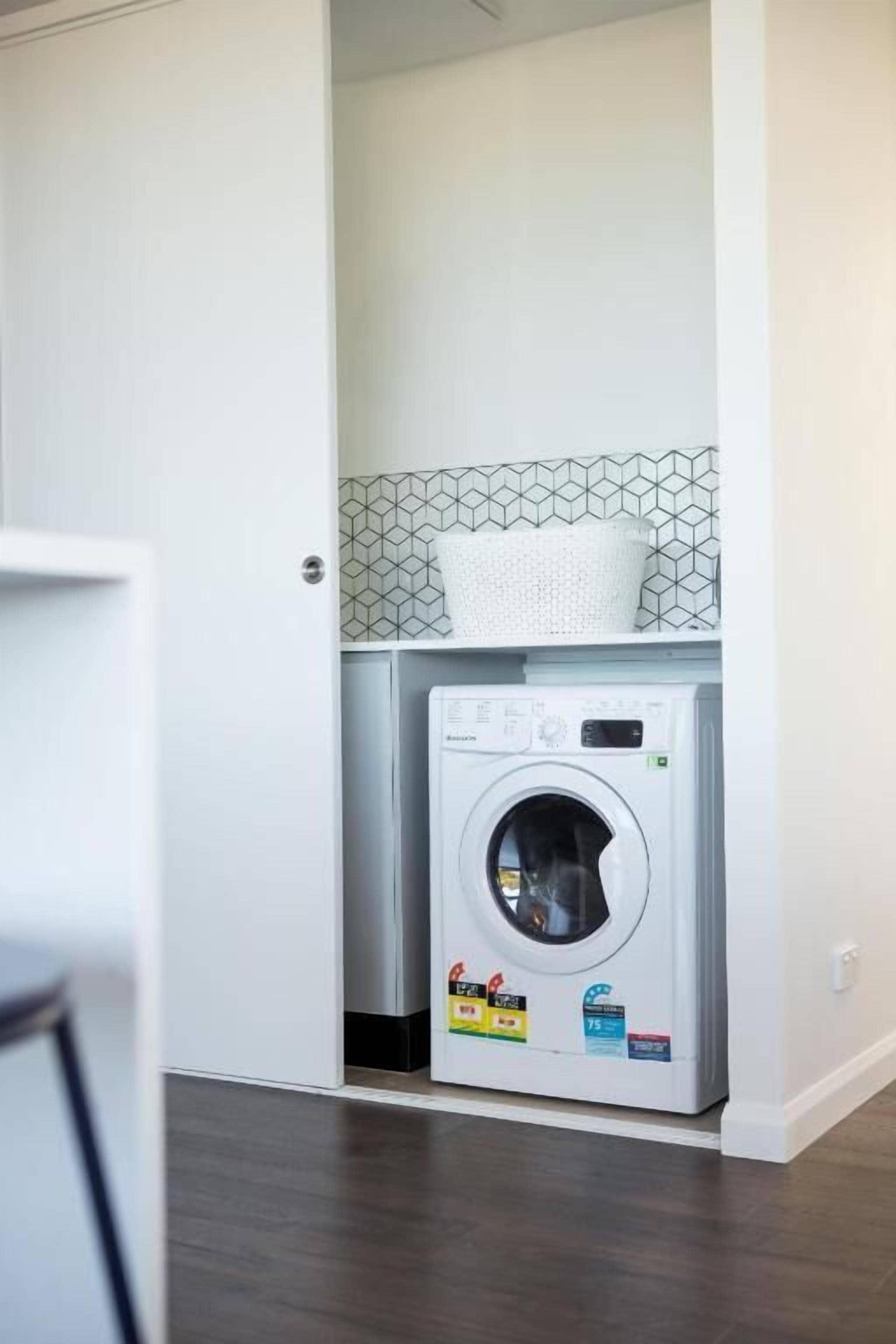 A compact laundry room with a front-loading washing machine, a tiled backsplash, and a basket on the shelf.