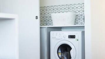 A compact laundry room with a front-loading washing machine, a tiled backsplash, and a basket on the shelf.