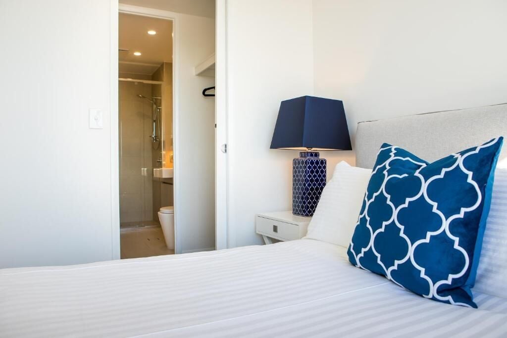 A hotel room with a bed, a nightstand, a lamp, and a bathroom visible through an open door.