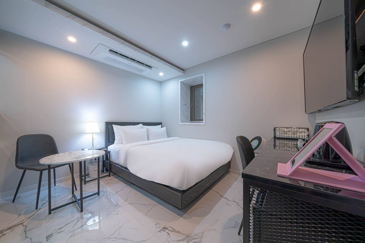 A modern hotel room with a bed, a desk, a chair, a lamp, and a TV.