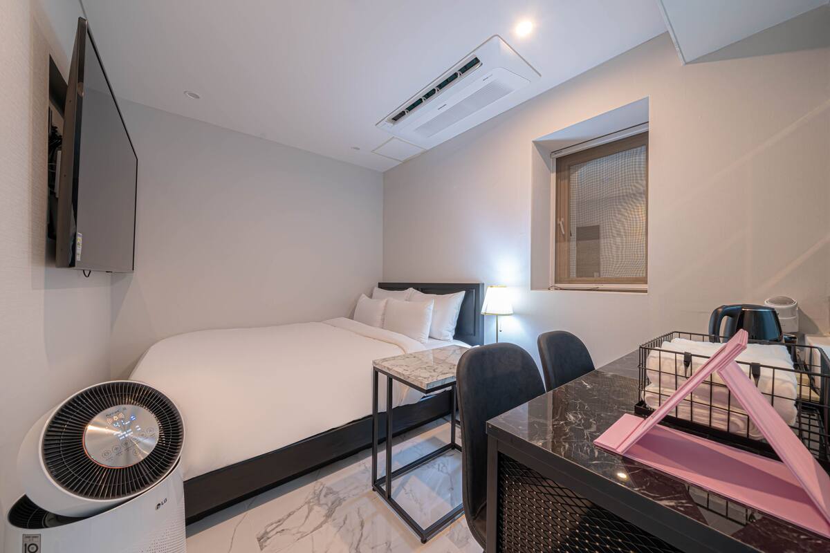 A hotel room with a bed, a desk with a chair, a standing fan, a TV mounted on the wall, and an air conditioning unit.