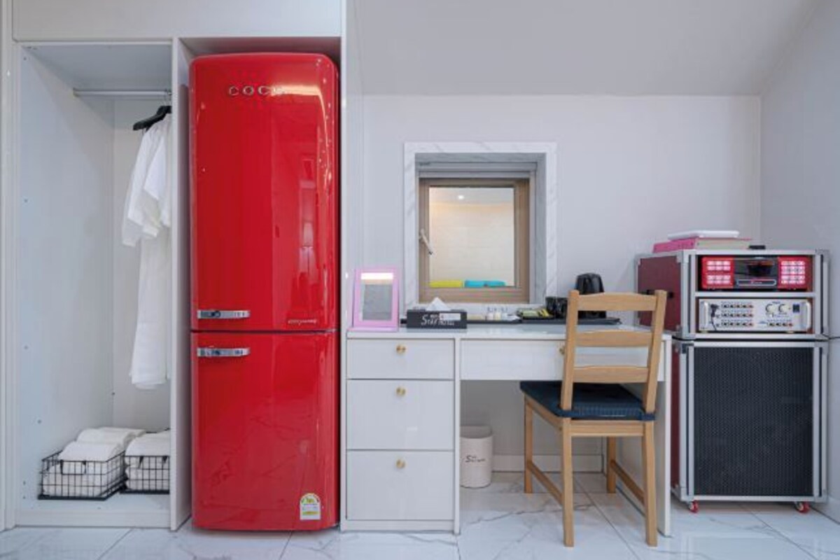 Une pièce de taille modeste, avec un réfrigérateur rouge, un petit bureau avec une chaise, un miroir et un haut-parleur.