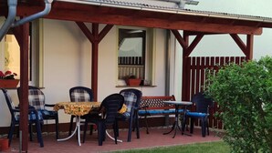 An outdoor patio with a table and chairs, a wooden pergola, a window with a curtain, and a small garden area.