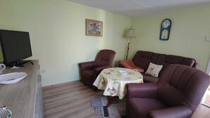 A living room with a brown leather sofa, a round coffee table with a floral tablecloth, a television on a wooden stand, and a clock on the wall.