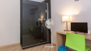 A room with a glass shower enclosure, a standing fan, a desk with a computer, a green chair, and a blue trash bin. - Hotel Genziana (Genoa)