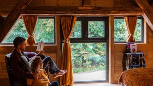 L’intérieur d’un chalet accueillant, avec des murs en bois, une grande fenêtre ornée de rideaux, une petite table avec une radio et un lit recouvert d’une couverture à motifs.