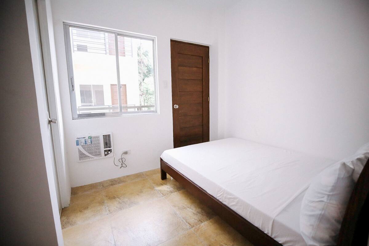 A bedroom with a bed, a window, a door, and a wall-mounted air conditioning unit.