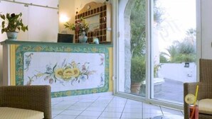 Uma área de recepção com piso de azulejos, balcão com padrão floral, móveis de vime e vista através de uma porta de vidro.