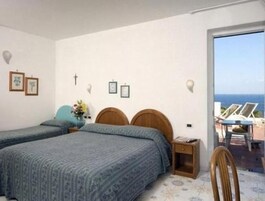 Quarto com duas camas, uma mesinha de cabeceira pequena, um criado-mudo e vista para o mar através de uma porta aberta.