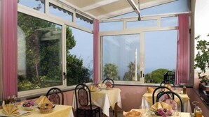 Uma sala de jantar com mesas preparadas para refeições, grandes janelas com cortinas e vista para o verde lá fora.